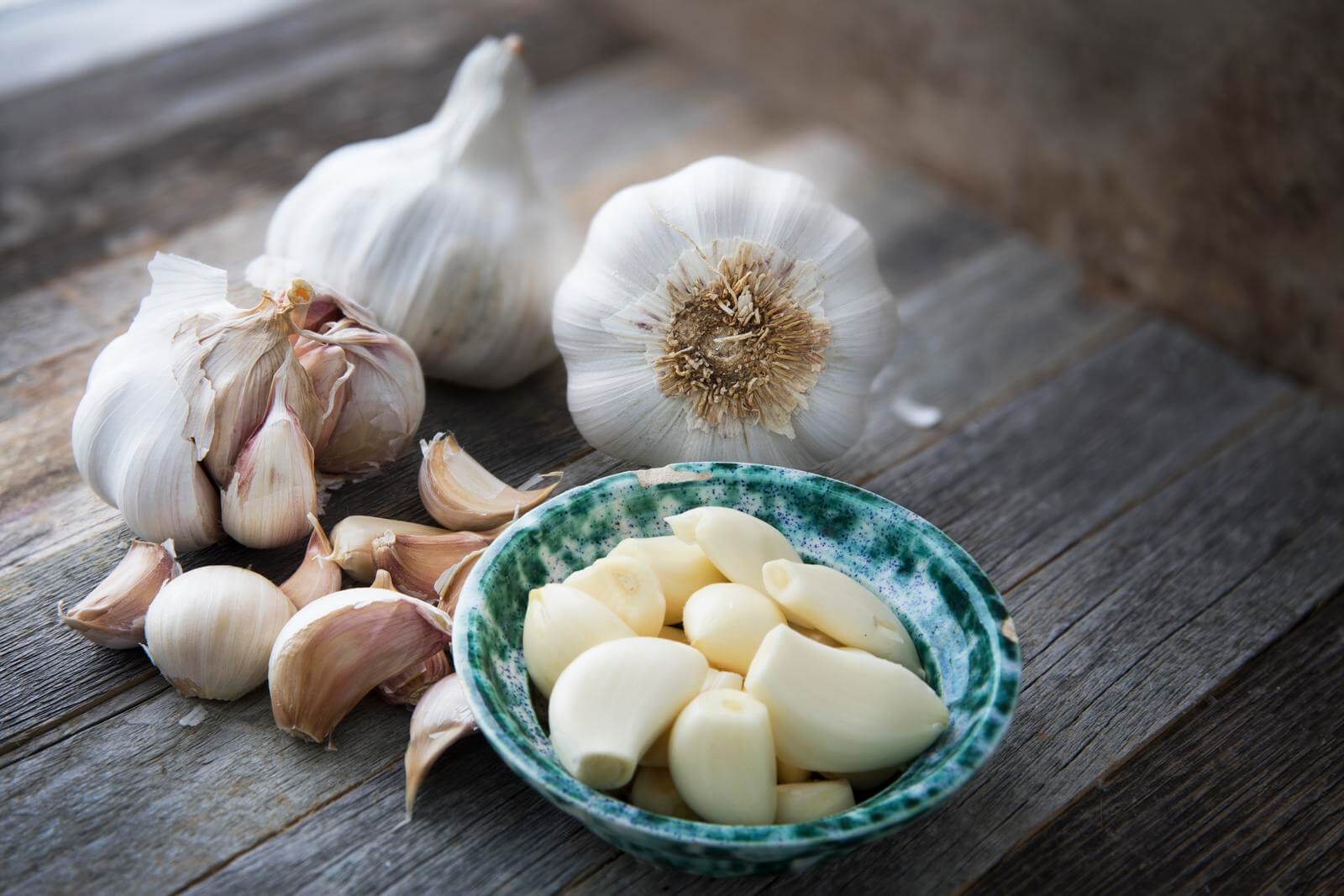 Bowl of peeled garlic cloves in bowl with more cloves and garlic heads on wooden surface