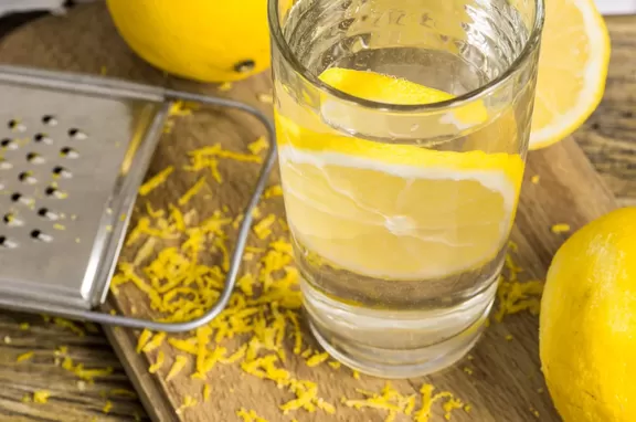 Glass of lemon water with lemon peel shavings on a wood board.