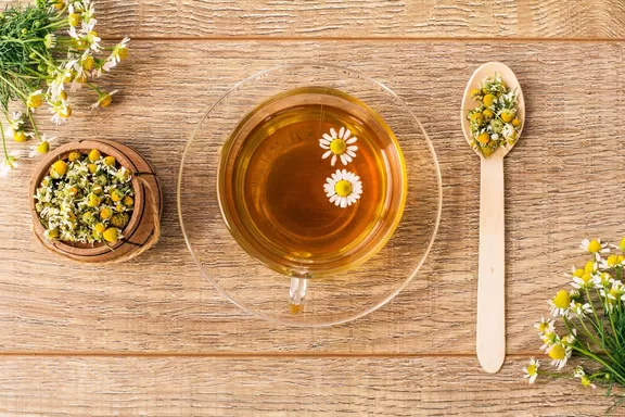 Glass cup of green tea with white chamomile flowers and little wooden barrel with dry flowers of chamomile on boards. Top view
