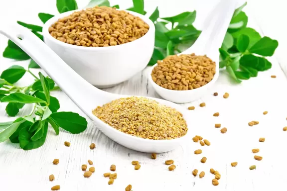 Fenugreek seeds and powder in two spoons and bowl with leaves on board.